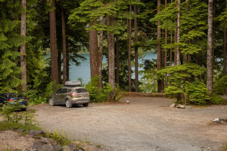 An SUV with a roof box is parked in at a Moutcha Bay Resort campsite surrounded by tall, green pine trees with a glimpse of a calm water visible through them.