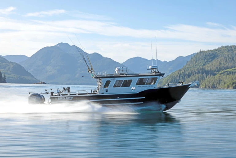 A sleek fishing boat with multiple rods speeds on calm water with scenic mountains and forests in the background on a clear day.